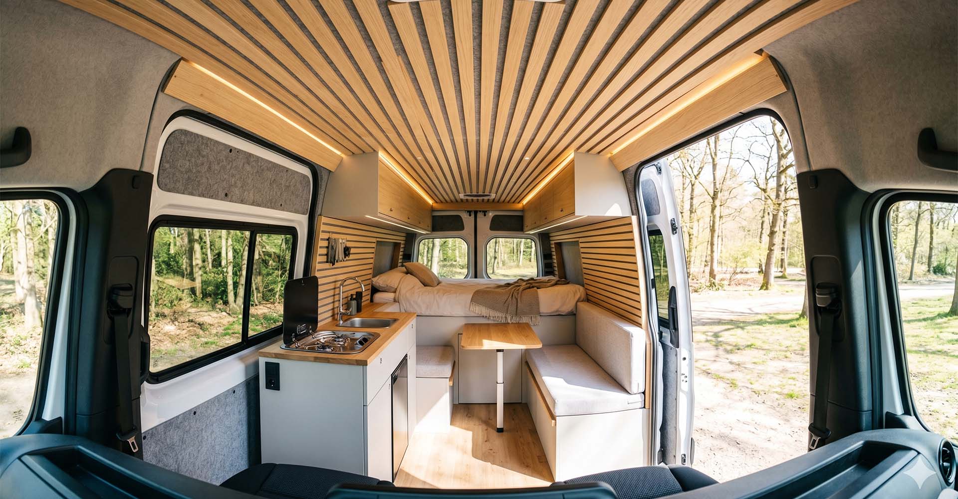 Interieur van een professioneel omgebouwde campervan met houten lattenplafond, ingebouwde keuken met spoelbak en gaskookplaat, zithoek met opklapbare tafel en een bed achterin, gefotografeerd vanuit de openslaande zijdeur met bosachtergrond.