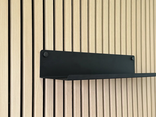 Zwarte stalen wandplank met opstaande voorrand op een eiken akoestisch wandpaneel, beide handschroeven in beeld