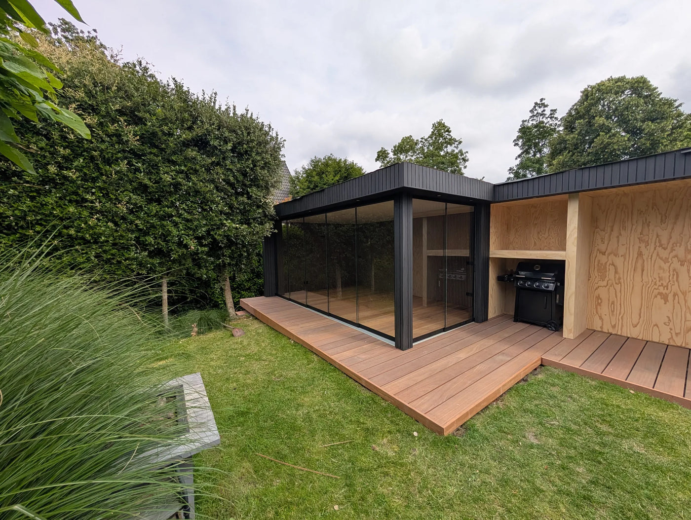 Moderne tuinkamer met zwarte gevelbekleding, glazen schuifpui en aangebouwde buitenkeuken op een hardhouten vlonder